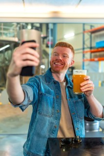 Smiling red haired man in a denim jacket takes a selfie with his smartphone while holding a glass
