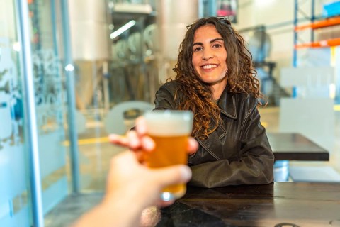 Woman smiling and raising a glass of frothy craft beer in a modern brewery, toasting an unseen