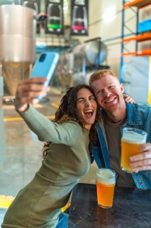 Happy young couple posing for a selfie with a smartphone, holding glasses of craft beer, and