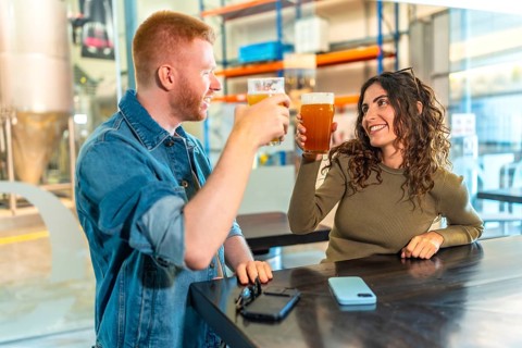 Happy young couple toasting craft beers and laughing together inside a modern brewery tasting room,
