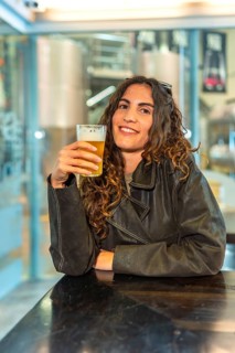 Smiling woman with curly hair wearing a leather jacket, happily holding a glass of pale beer while