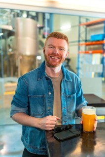 Smiling man enjoying a glass of craft beer in a modern industrial brewery. He stands wearing a