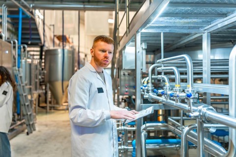 Young male specialist wearing a white lab coat and observing pipes and machinery while holding a