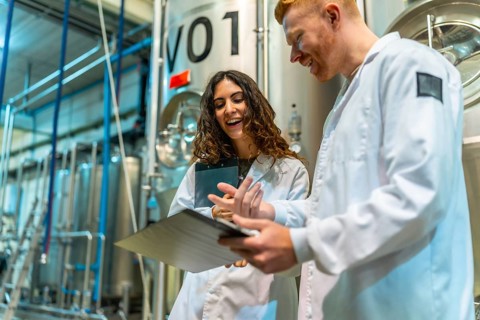 Brewery colleagues smiling and discussing production notes on a clipboard, standing in a beer
