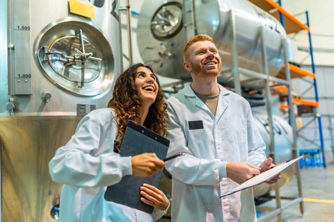 Smiling man and woman in lab coats holding clipboards, overseeing production and quality control in