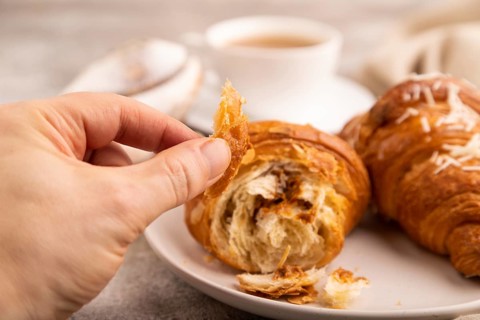 Croissant on white plate with hand on brown concrete background and linen textile, cup of coffee,