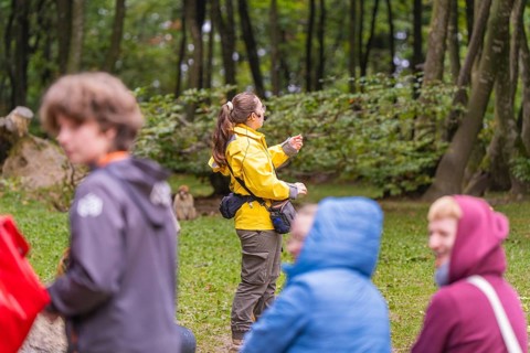 A person in a yellow jacket points to something in a wooded area. Surrounded by blurred figures,