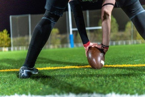 American football player in dark uniform with arm sleeves placing a brown ball on green turf at