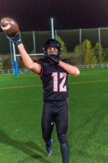 American football player wearing a black uniform with number 12 and helmet, extending arm to catch
