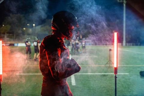 American football player standing on a dark field at night in helmet and pads, framed by red and