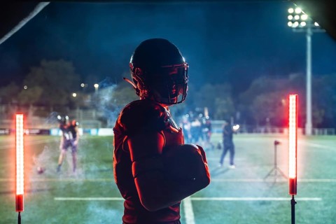 American football player in full gear holding a ball, standing confidently on a floodlit stadium