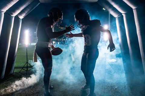 American football players, one holding a ball, emerging from a smoky stadium tunnel at night,