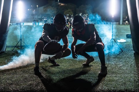 Two american football players in full gear, crouching and ready for action, emerging from a dark
