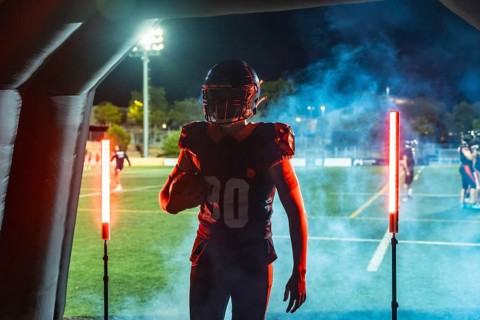 American football player in helmet and uniform bursts from a smoke filled tunnel onto a floodlit