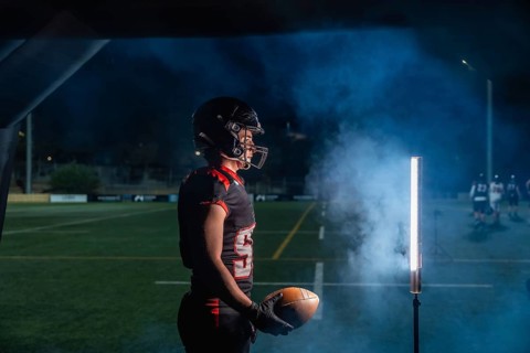 American football player is holding a ball, wearing a helmet and uniform, and standing on a playing