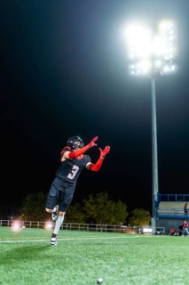 American football player in uniform with a number 3 catching a ball on a lit artificial turf field