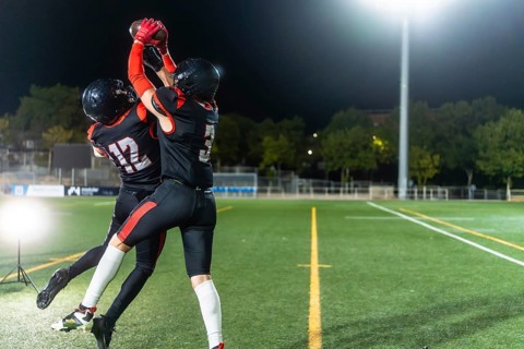 Two female american football players in black uniforms leap to catch the ball during a night game
