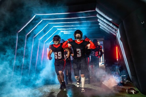 American football players in black and red uniforms and helmets running through a dramatic, smoke