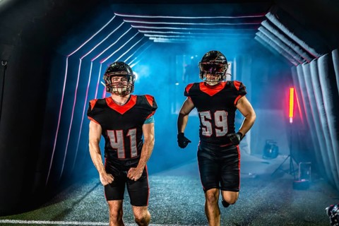 American football players in uniforms and helmets are stepping out from a dimly lit tunnel onto a