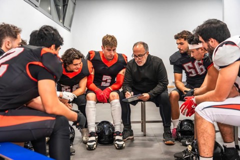 American football coach discussing strategy with his team of male players in the changing room,