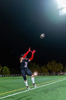 American football player in uniform and helmet is jumping to catch a football during a night game