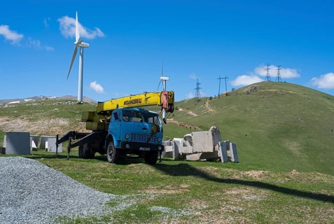 A blue crane lorry stands on a green construction site next to a wind turbine on a hill, Pushkin