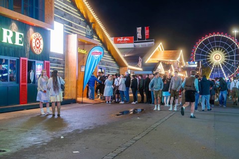 Crowd in front of illuminated festival tents and a large Ferris wheel at night, Cannstatter Wasen