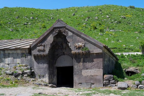 Old stone building with detailed decorations in front of a lush green hill on a sunny day,