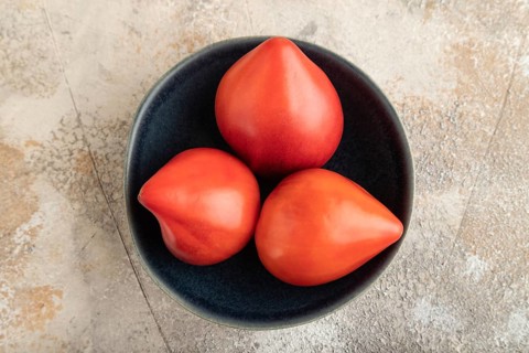 Red Heart shape tomatoes in blue bowl on brown concrete background. Top view, copy space, flat lay.