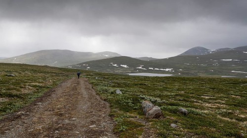 Hiker with rucksack and blue raincoat on a lonely plateau with snowfields, low clouds, Kongevegen