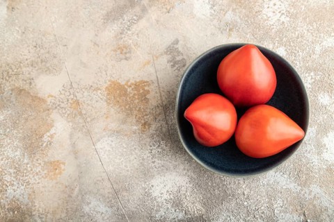 Red Heart shape tomatoes in blue bowl on brown concrete background. Top view, copy space, flat lay.