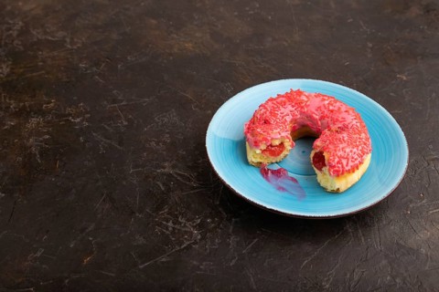 Bitten Pink Donut with sprinkles on blue ceramic plate on black concrete background, side view,