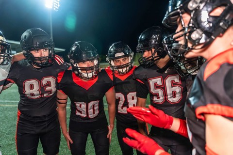 American football players huddling on a lit turf field at night, helmets on, focused on strategy