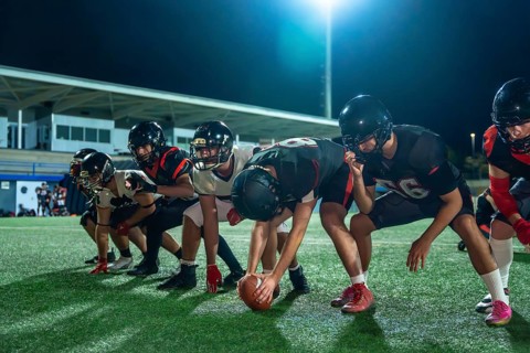 American football players in full uniform and helmets lining up on the green grass field at night,