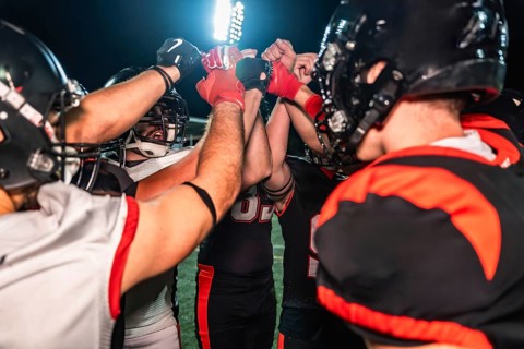 American football players huddling on the illuminated field, placing their hands together in the