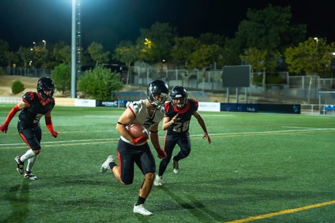 American football player vigorously running with the ball on a green field during a night game, two