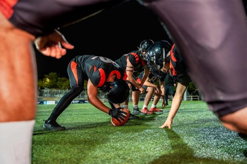American football players line up on the field at night, a center snapping the ball, preparing for