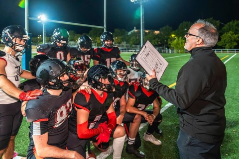 American football coach holds clipboard with play diagram, instructing young athletes in full