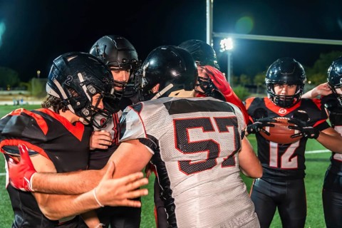 American football players in black and red uniforms with helmets and shoulder pads huddling on a