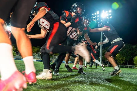 American football players in black and red uniform helmets battling on a green artificial turf