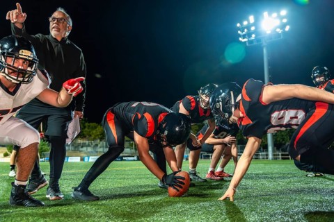 American football team huddled on a dark stadium field under bright lights at night, coach pointing