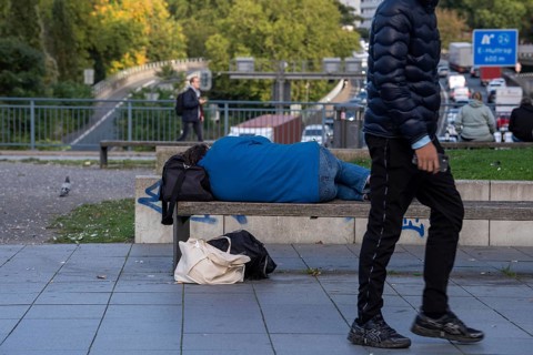 Man sleeping on a bench, travelling bag serves as pillow, in the city centre, passers-by walk by