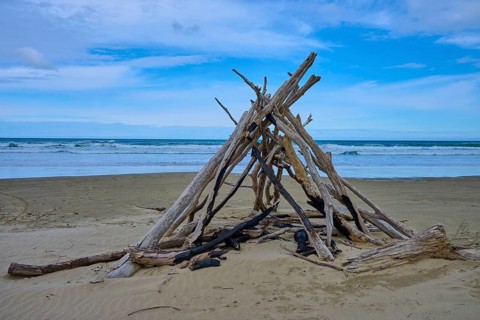 Treibholzstruktur in Zeltform am Strand unter blauem Himmel und Meerblick, Kaka Point, Clutha