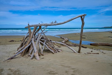 Treibholzskulptur Zelt am Strand unter blauem Himmel, ruhiges Meer im Hintergrund, Kaka Point,