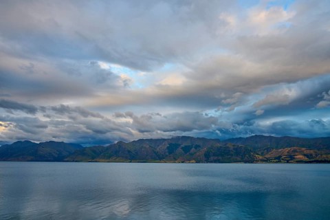 Stilles Gewässer vor bergiger Landschaft und dramatischem Himmel in der Dämmerung, Lake Hawea,