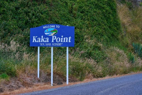 Willkommensschild von Kaka Point neben einer Straße mit grüner Vegetation und landschaftlicher