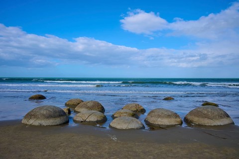 Runde Felsen verteilen sich am Strand unter einem blauen Himmel mit Wolken, Moeraki Boulders,