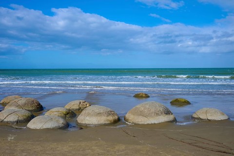 Mehrere runde Felsen an einem Sandstrand mit Blick auf das Meer und bewölkten Himmel, Moeraki