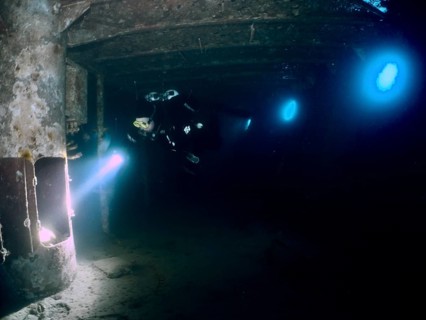Diver exploring a dark wreck underwater, illuminated by torchlight. Dive site wreck of the Vis,