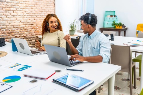 Two smiling multiracial coworkers wearing headsets, collaborating and working on laptops at a